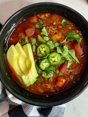 A bowl of cooked Slow Cooker Venison Chili topped with sliced avocado, chopped cilantro and sliced jalapenos.