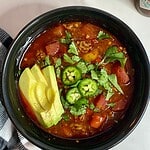 A bowl of cooked Slow Cooker Venison Chili topped with sliced avocado, chopped cilantro and sliced jalapenos.