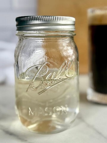 A jar of Sugar-Free Simple Syrup with a cup of coffee in the background.